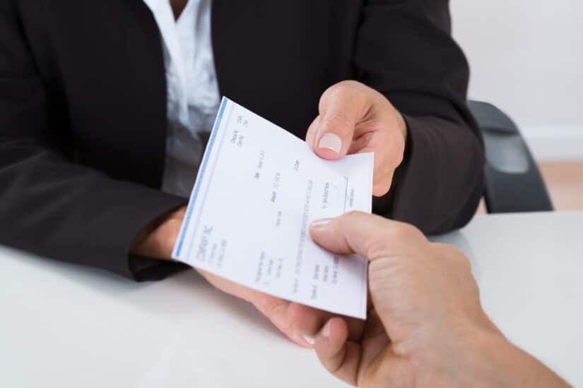 Close-up Of Businessperson Hands Giving Cheque To Other Person At Desk