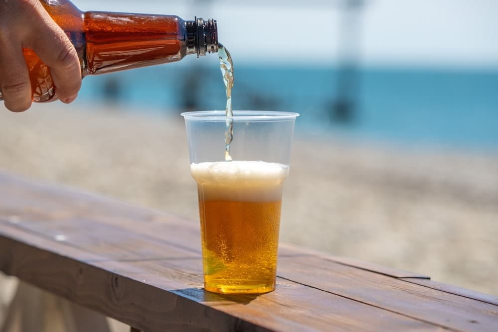 Open Beer Container A person is pouring beer into a plastic cup on a wooden bench. The scene is relaxed and casual, with the beach setting adding to the laid-back atmosphere